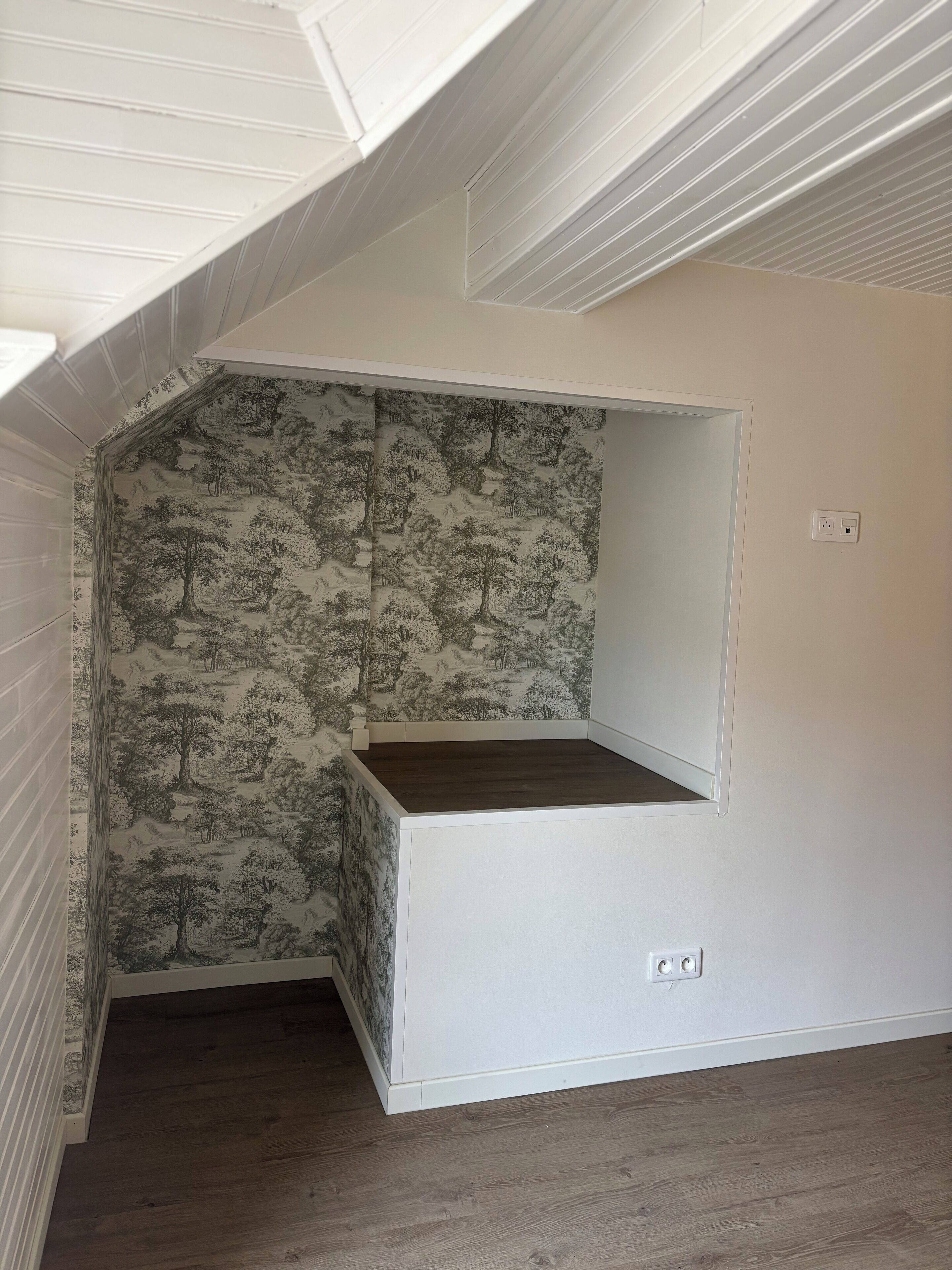 A room with a sloped ceiling, a built-in bench, and wallpaper with a tree pattern.