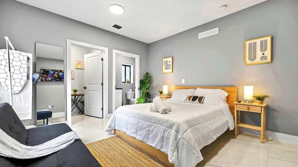 Quarto moderno com cama grande, mesa de cabeceira com abajur, cadeira e TV fixada na parede.