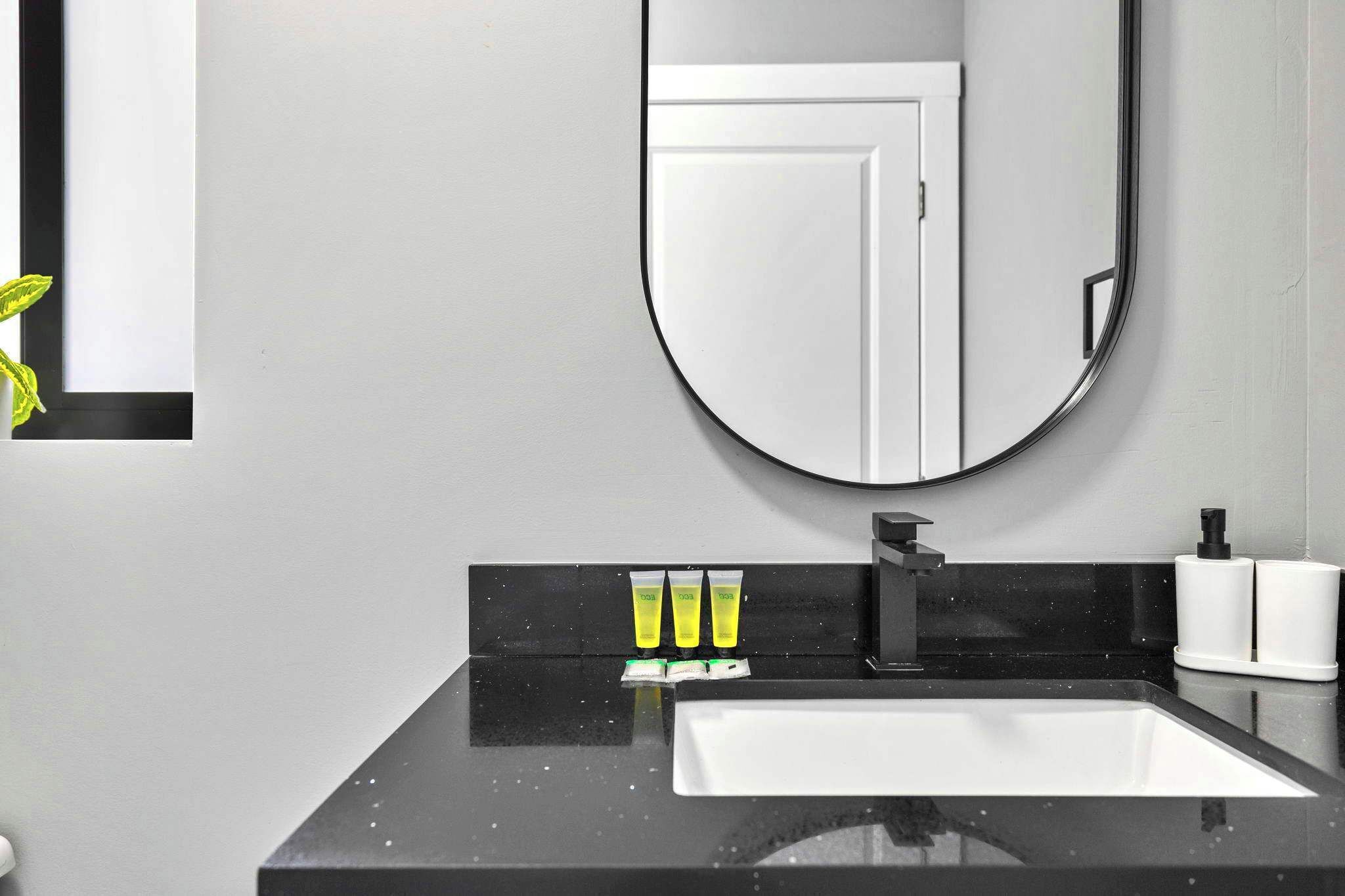 Une salle de bain moderne avec un plan de toilette noir, un lavabo blanc et un miroir rond. On peut voir trois tubes jaunes sur le plan de toilette, et une porte blanche se reflète dans le miroir.