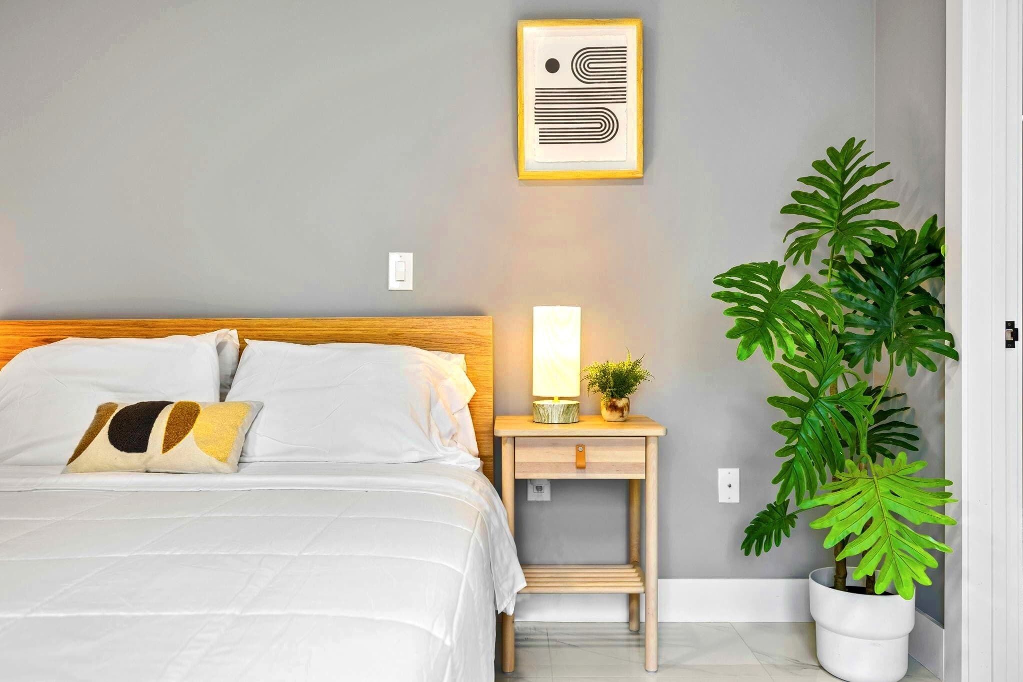Una cama bien hecha con ropa blanca y almohadas estampadas, una mesita de noche con una lámpara y una planta pequeña, y una planta verde grande en una maceta blanca contra una pared gris.