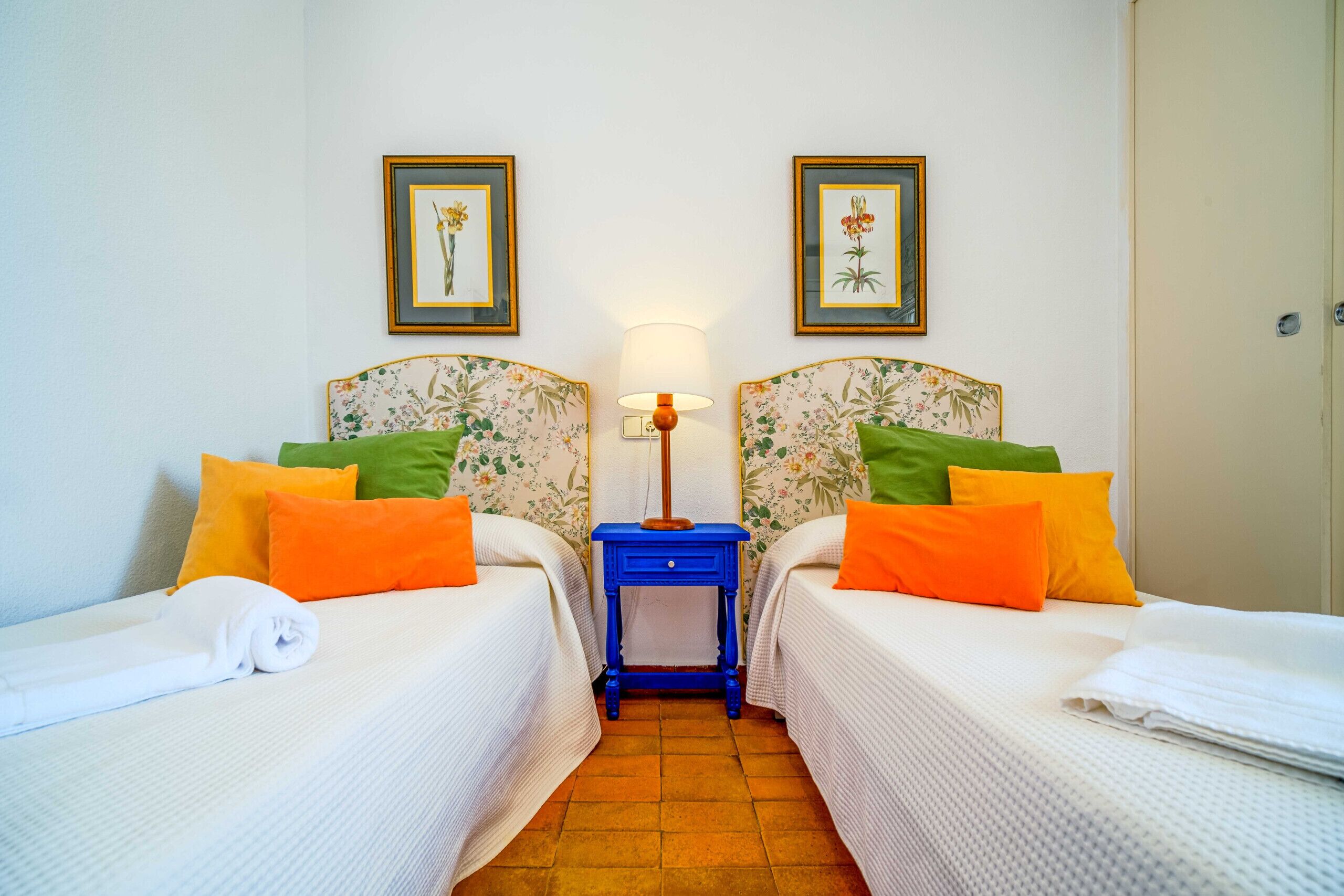 Une chambre d’hôtel avec deux lits, chacun doté d’une tête de lit à motifs floraux, une petite table de chevet avec une lampe, et deux tableaux encadrés accrochés au mur.
