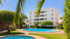 Un área de piscina con dos piscinas, rodeada de abundante vegetación y un edificio de varios pisos al fondo.