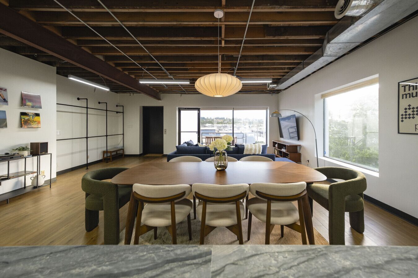 A modern dining area with a wooden table and chairs, a hanging light fixture, and a view of the outdoors through large windows.
