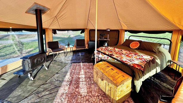 Una tienda de campaña con una cama, una mesa, sillas, una estufa y vistas al exterior.