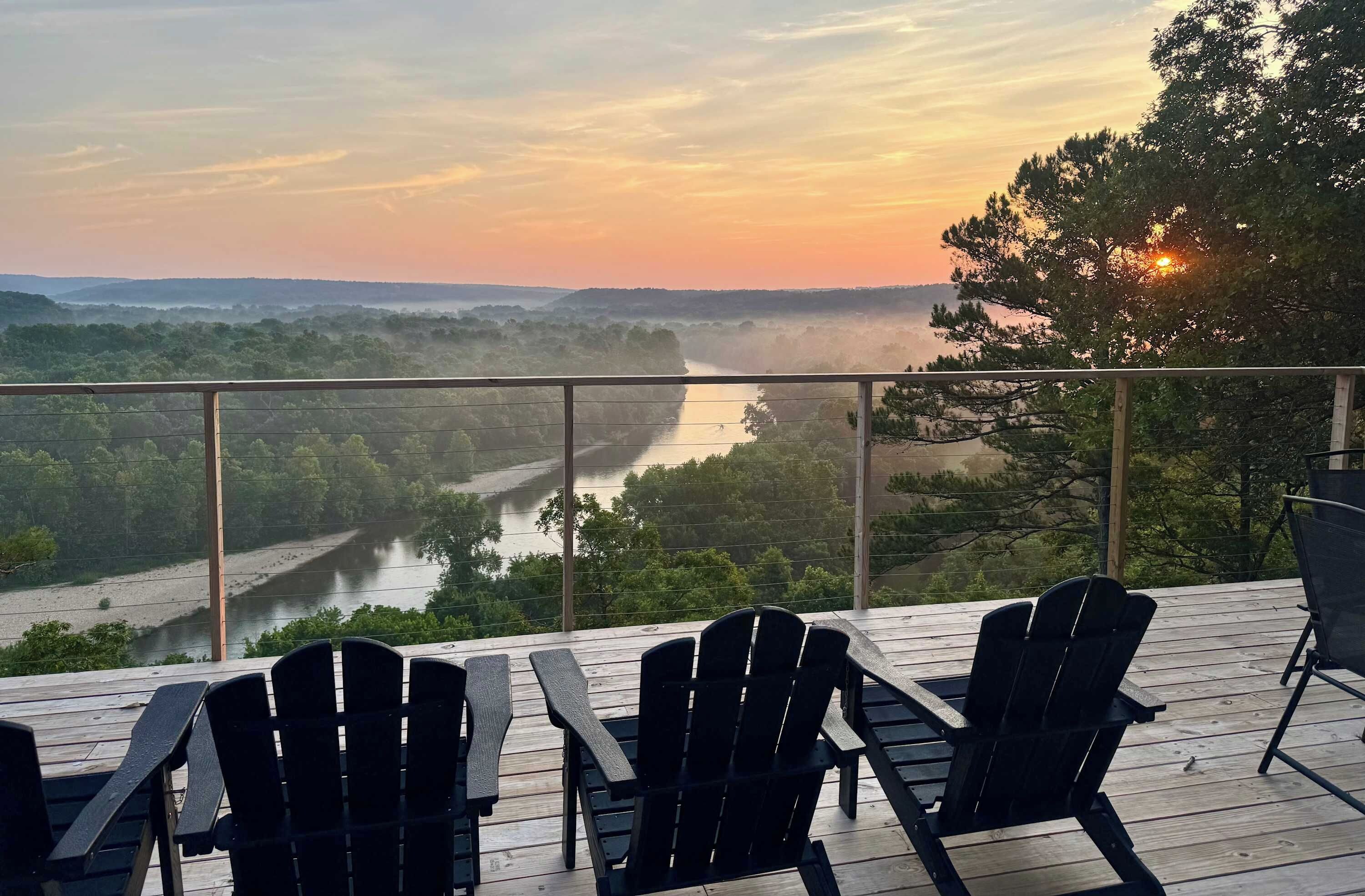 A deck with Adirondack chairs overlooking a river and forested landscape during sunset.