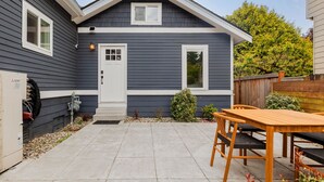 Ett bostadshus med en grå fasad, en ytterdörr, en liten veranda och en uteplats med ett träbord och stolar.