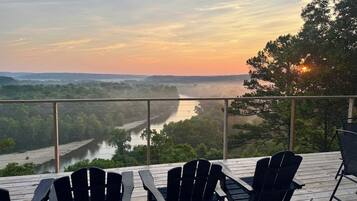 A deck with Adirondack chairs overlooking a river and forested landscape during sunset.
