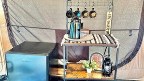Una kitchenette compacta con un refrigerador pequeño, un estante metálico con ollas y sartenes, un farol y una pequeña planta en maceta.