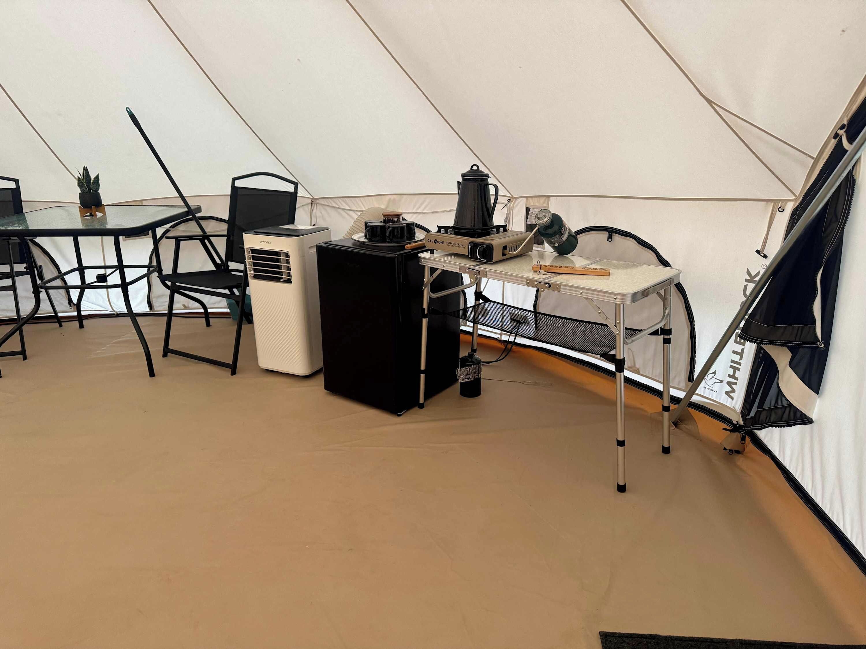 Ein Zeltinnenraum mit Tisch, Stühlen, einem tragbaren Heizgerät, einem Ventilator und einer Kaffeemaschine.