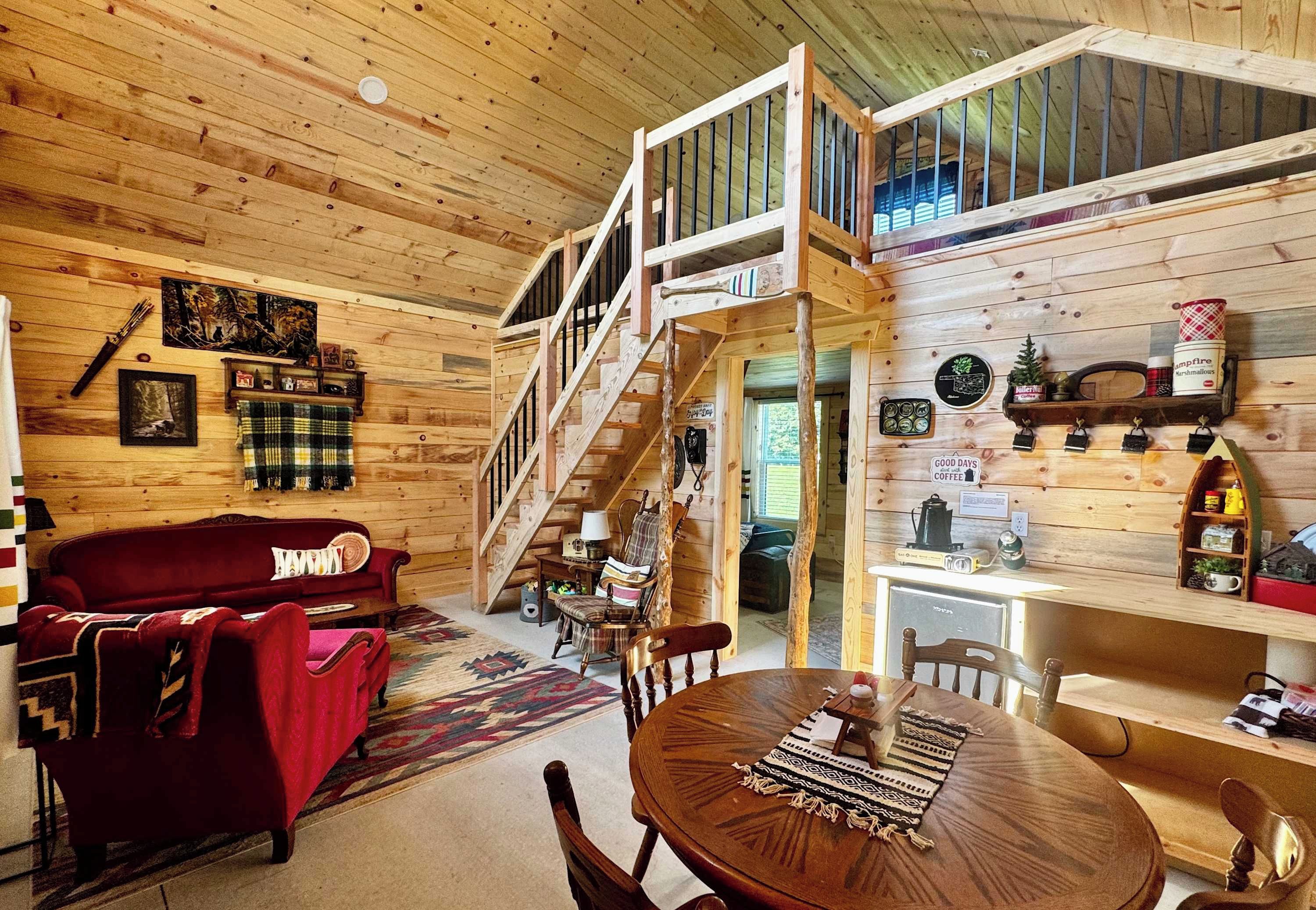 A wooden cabin interior with a red sofa, a dining table, a staircase, and various decorations.