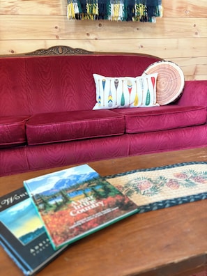 A red sofa with decorative pillows, a wooden side table with magazines, and a wooden wall with a hanging tapestry.