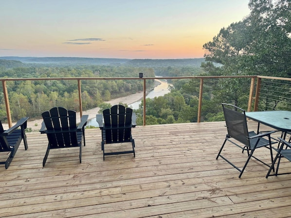A deck with chairs and a table overlooking a river and forested landscape.