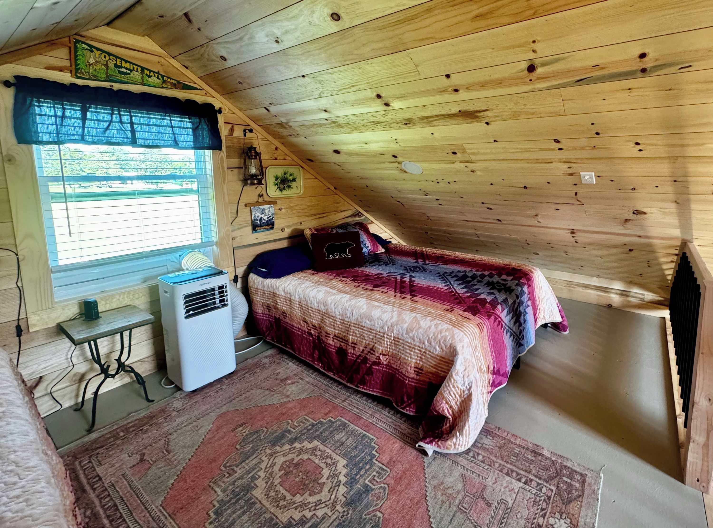 Ein gemütliches Schlafzimmer im Dachgeschoss mit schrägen Wänden, einem Bett mit gemustertem Überwurf, einem kleinen Tisch und einer Klimaanlage sowie einem Fenster mit Blick ins Freie.