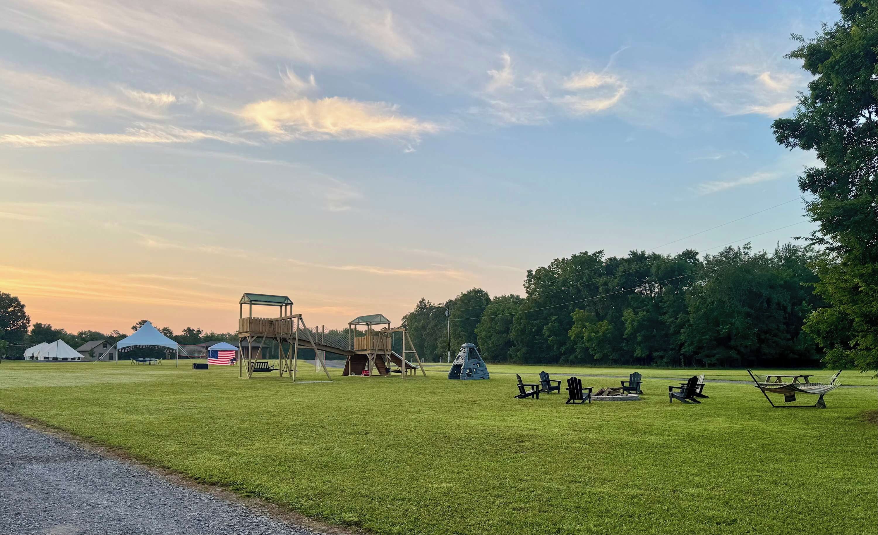 Eine grüne Wiese mit einem Spielplatz, Picknicktischen und Adirondack-Stühlen.