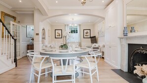 A dining room with a round table, white chairs, a chandelier, framed artwork, and a fireplace. - Gorgeous House - Garden & Open Living (Cheltenham)