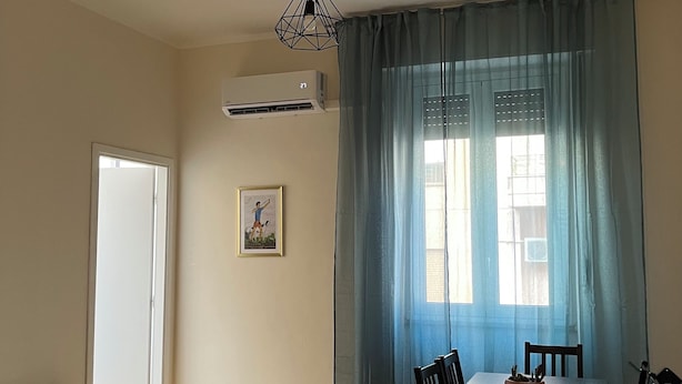 Habitación de hotel con cama, mesa de comedor con sillas, lámpara de techo, unidad de aire acondicionado y ventana con cortinas.