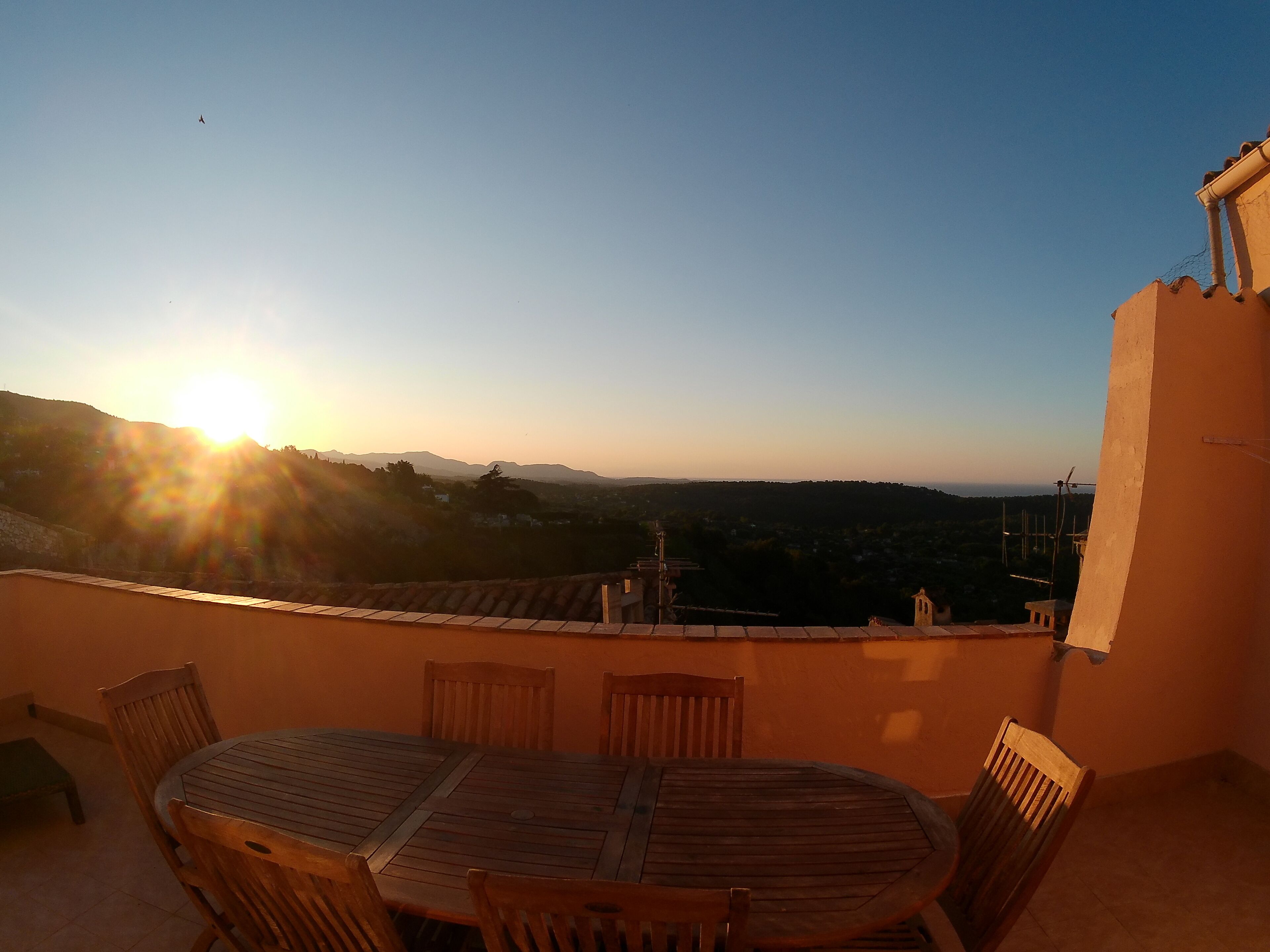 Une table et des chaises en bois sur une terrasse extérieure, offrant une vue sur le paysage et les collines lointaines au coucher du soleil.