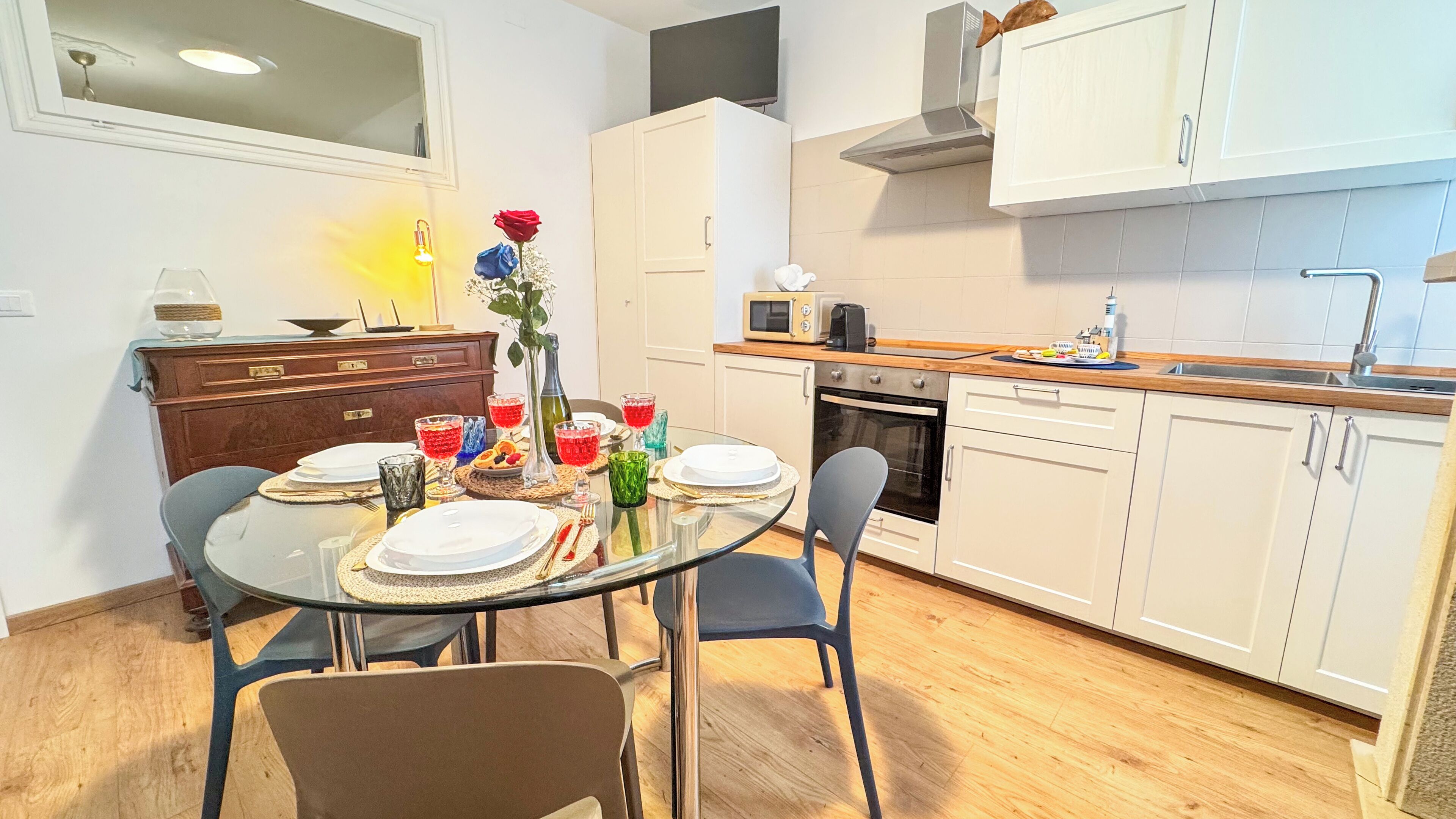 Une cuisine moderne avec des meubles blancs, un four à micro-ondes et un évier. On y trouve une table à manger mise avec des assiettes, des verres et un vase rempli de fleurs.