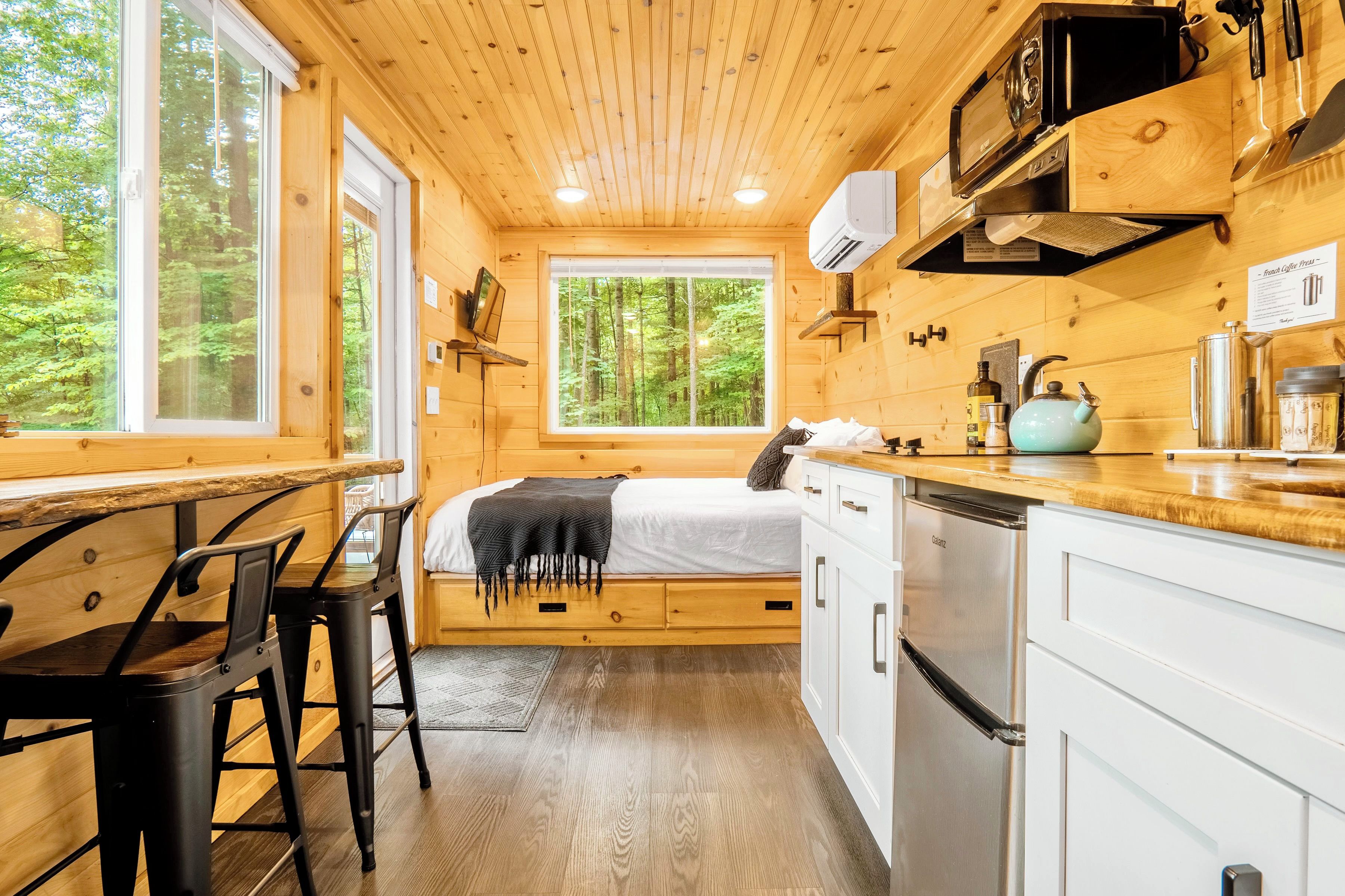 L’intérieur d’une cabane comprend un lit, des murs en bois, un coin cuisine et une fenêtre donnant sur une forêt.