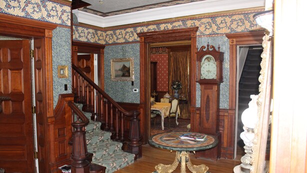 A grand hallway with a wooden staircase, a round table with a book, and a clock on the wall.