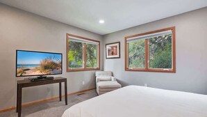 A bedroom with a bed, a television on a table, and two windows with a view of greenery.