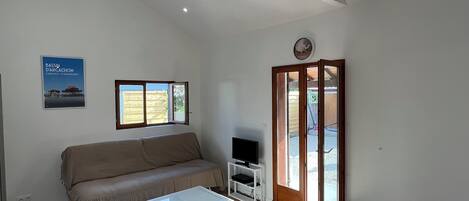 A room with a sofa, a coffee table, a TV on a stand, a dining table with a bowl, a window, and a door leading outside.