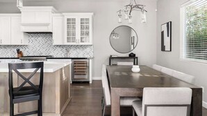 Een moderne keuken met witte kastjes, een backsplash in een herringbone patroon, een centraal eiland met een ingebouwde oven, en een eethoek met een houten tafel en stoelen.