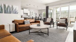 Een moderne woonkamer met een bank, salontafel en stoelen, een keuken op de achtergrond, en een grote schuifglazen deur die leidt naar een balkon.