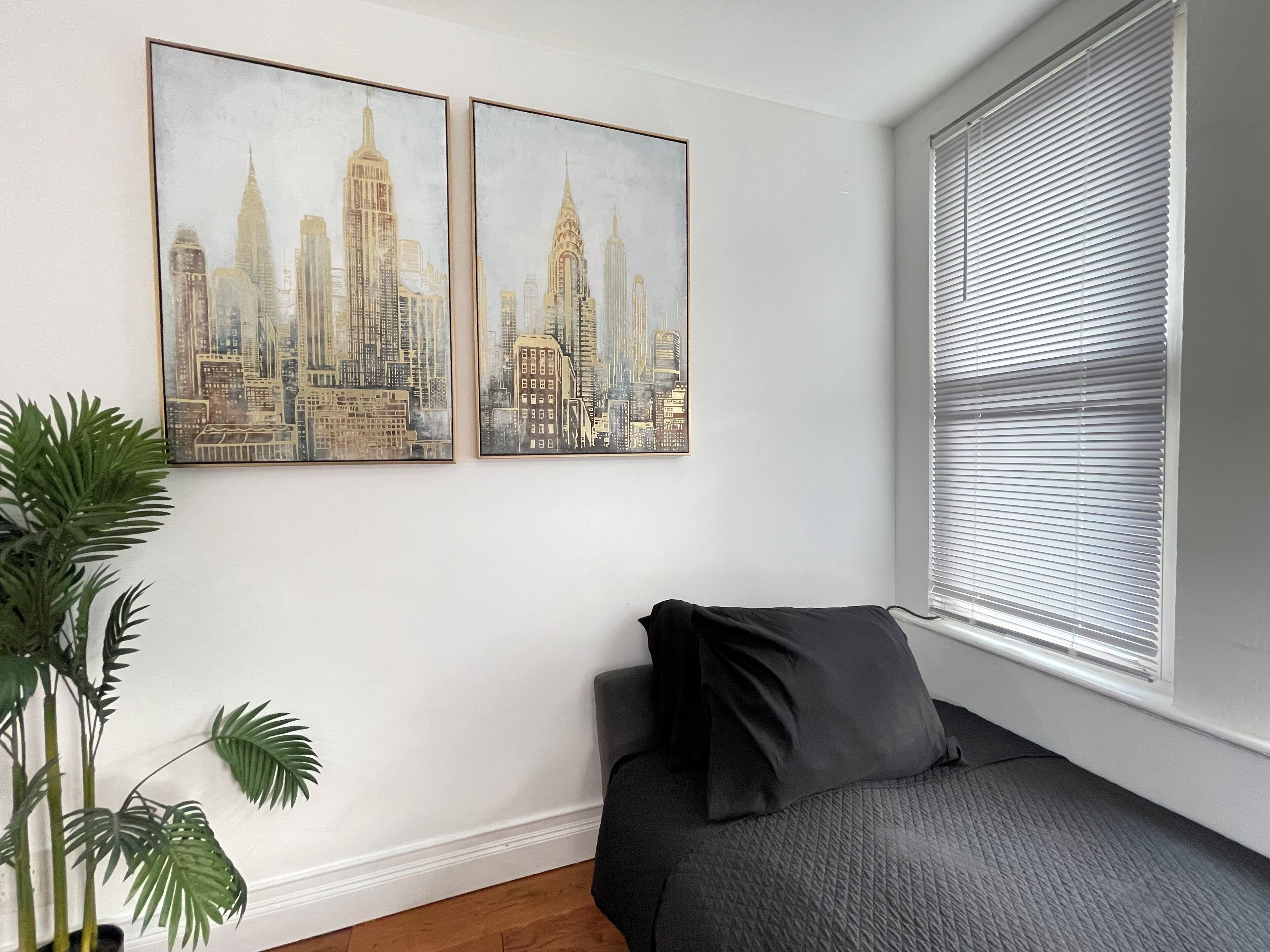 Une chambre à coucher avec un lit, deux tableaux représentant des paysages urbains, une fenêtre avec des stores et une plante en pot.