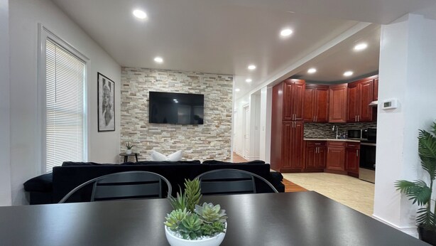 Un espacio habitable moderno con comedor, destacando una mesa negra y un cuenco blanco con suculentas. Un televisor de pantalla plana está instalado en una pared con textura de piedra, y la cocina cuenta con gabinetes de madera y un microondas.