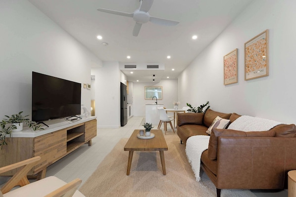 Uma sala de estar moderna com televisão de tela plana, mesa de centro de madeira, sofá de couro marrom e um espaço de jantar com mesa e cadeiras brancas.