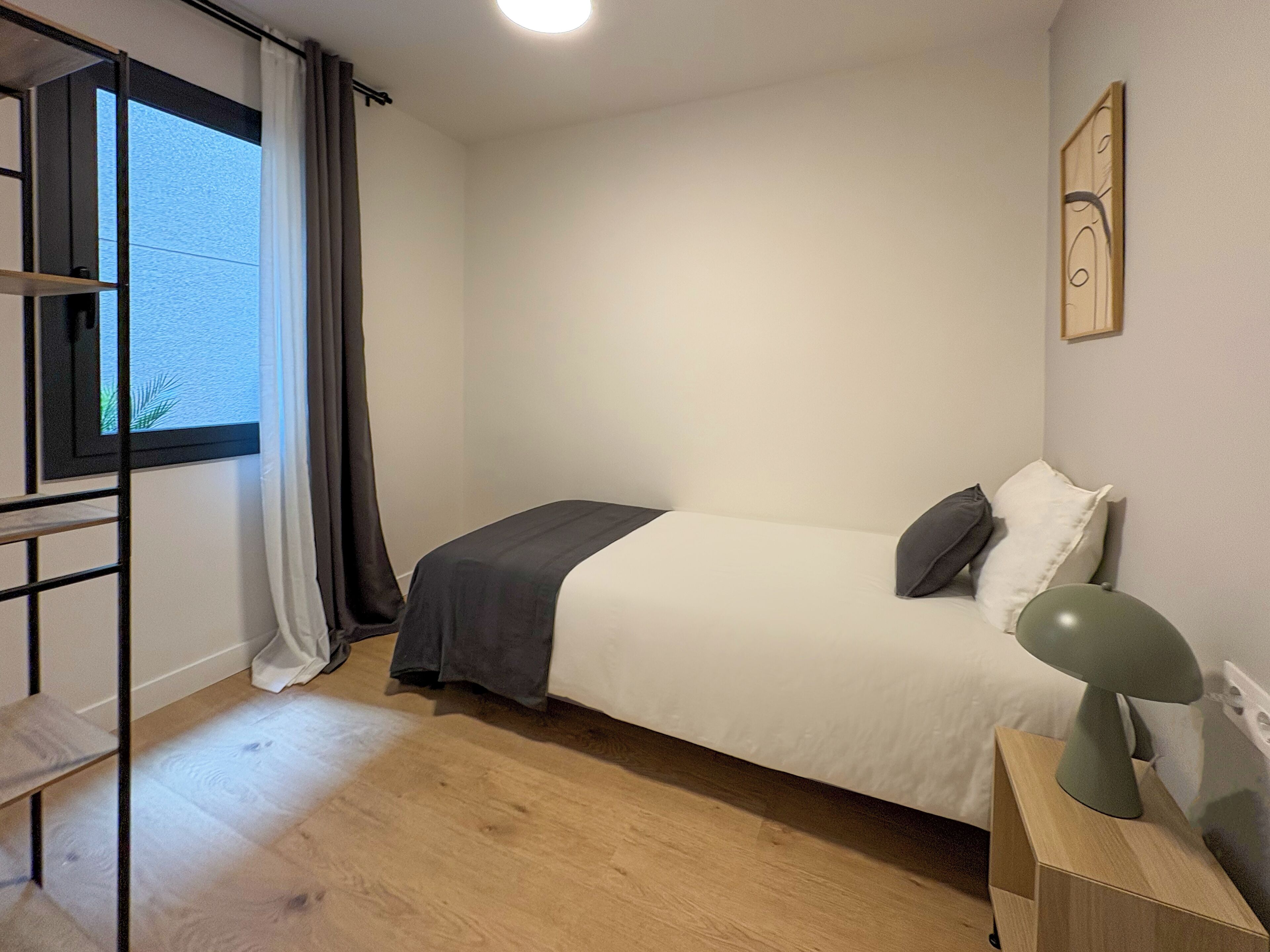 A bedroom with a bed, a bedside table, a lamp, a window with curtains, and a wall-mounted artwork.