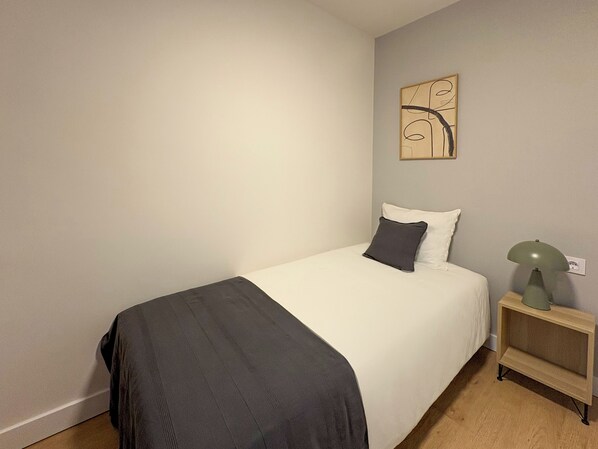 A neatly made bed with a white and dark grey pillow, a grey blanket, and a small bedside table with a lamp.