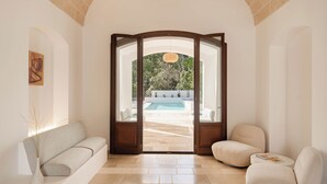 A modern interior with a sofa, a chair, and a small table. There is a door leading to an outdoor area with a pool. - Luxury villa with private pool | Villa Arabesca (Selva di Fasano)