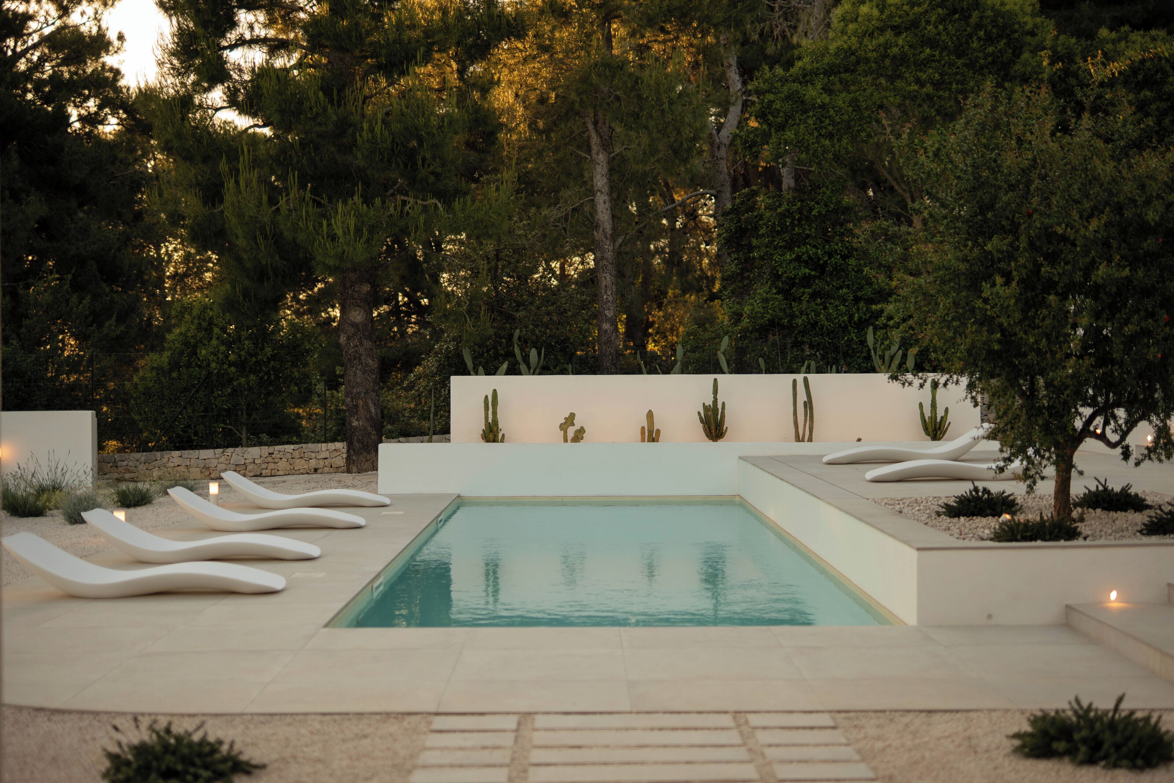 A rectangular pool with white loungers, surrounded by cacti and trees, with a stone wall and a white building in the background.