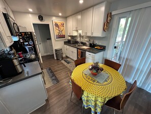 A modern kitchen with white cabinets, a dining table with a yellow tablecloth, and a window with blinds. - 2-bedroom condo in welcoming Terra Linda. Serene & convenient, fully equipped. (San Rafael)
