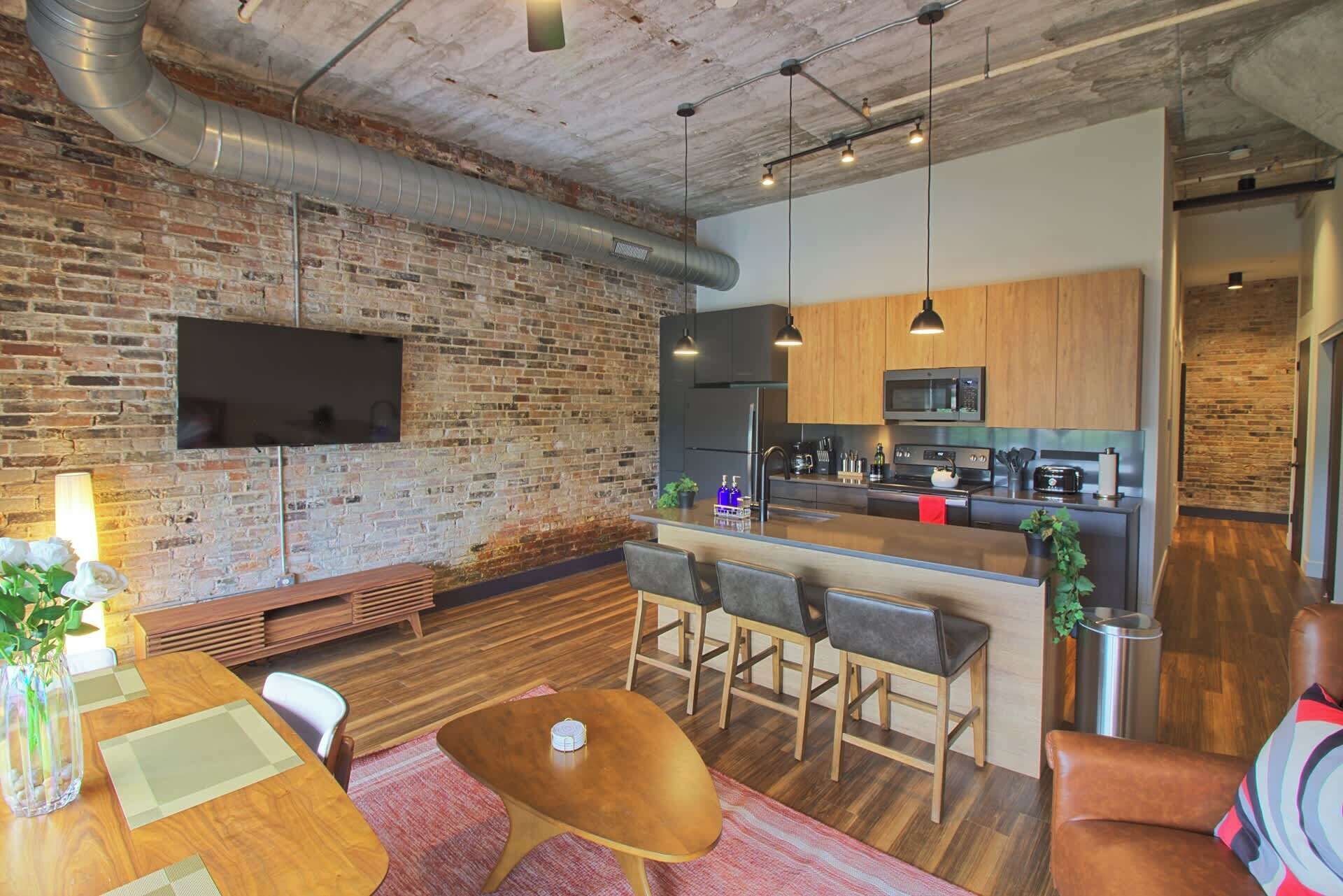 A modern kitchen with a central island, bar stools, and a flat-screen TV mounted on a brick wall.