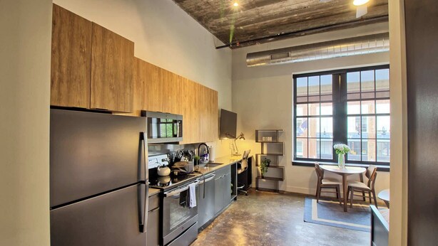 A modern kitchen with stainless steel appliances, wooden cabinets, and a large window offering a view of the cityscape. - Chic Ultramodern Studio Loft w/ Designer Features (St. Louis)