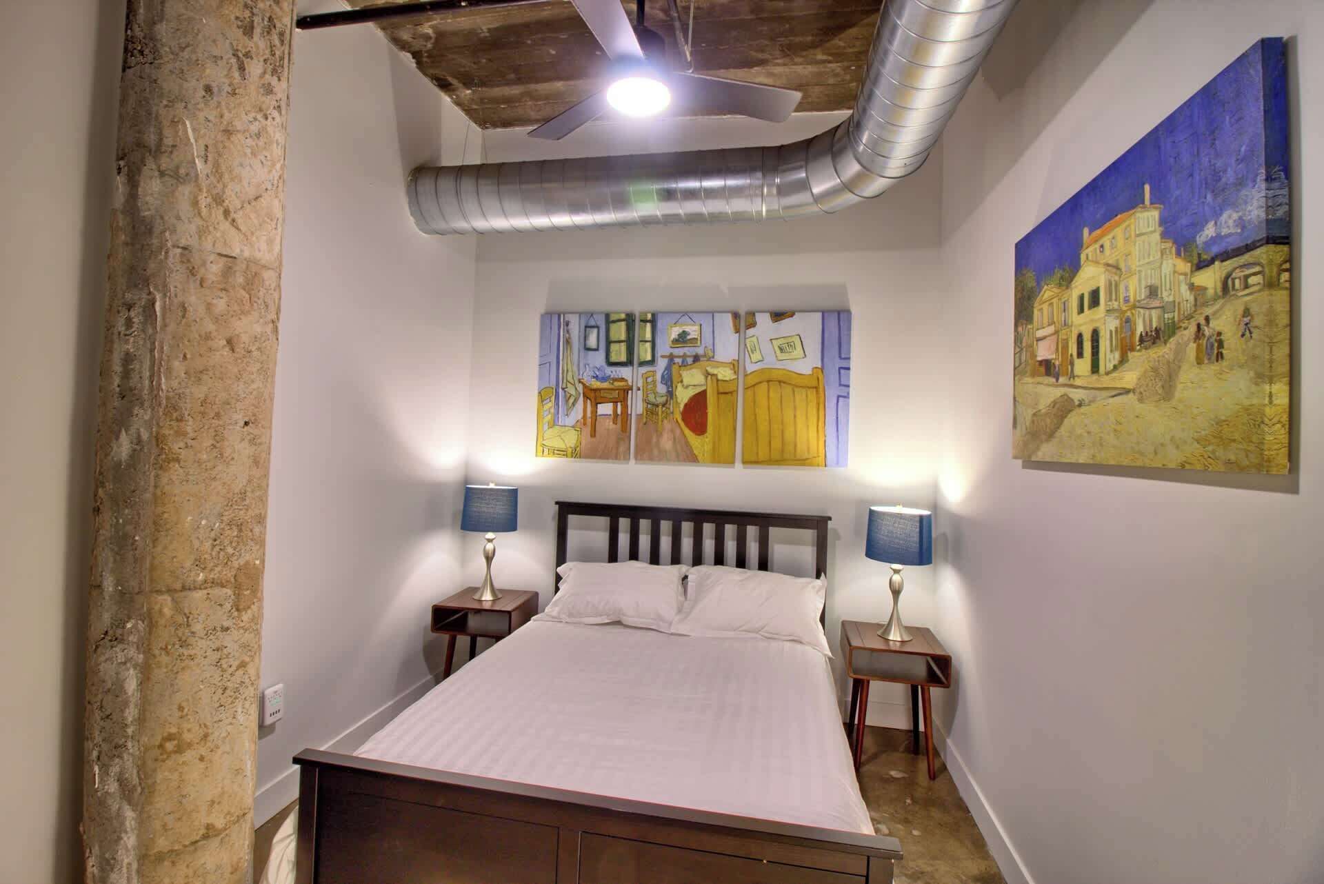 A bedroom with a bed, two bedside tables with lamps, a ceiling fan, and two paintings on the wall.