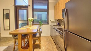 A modern kitchen with a stainless steel refrigerator, wooden cabinets, a dining table with chairs, and large windows.