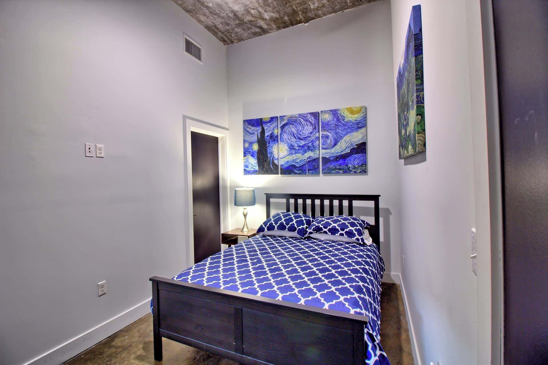 A bedroom with a bed, a nightstand, a lamp, and three paintings on the wall.