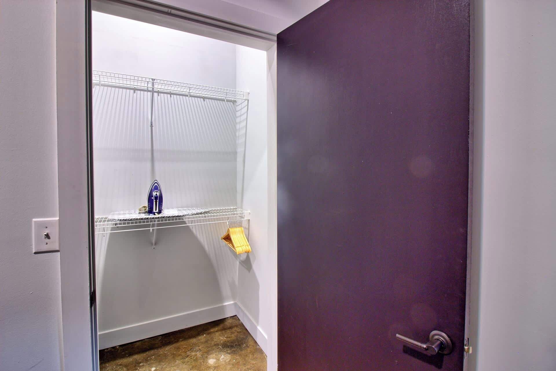 Una habitación con una puerta morada, una pared blanca y una estantería metálica con un planchado azul y un objeto amarillo.