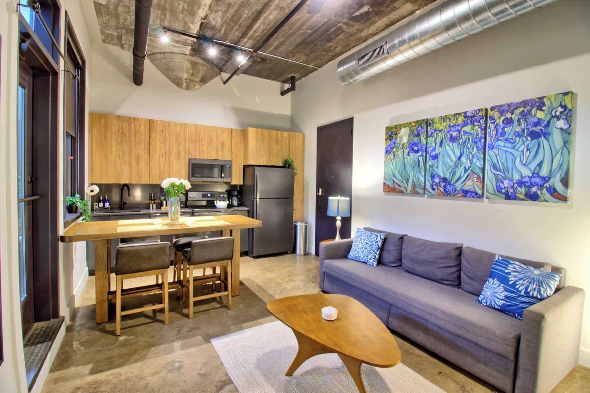 A modern kitchen with wooden cabinets, a dining table with chairs, a refrigerator, and a microwave. There is a gray sofa with patterned cushions and a coffee table.