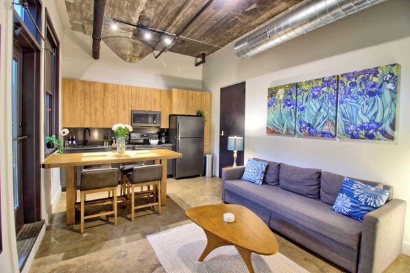 A modern kitchen with wooden cabinets, a dining table with chairs, a refrigerator, and a microwave. There is a gray sofa with patterned cushions and a coffee table.