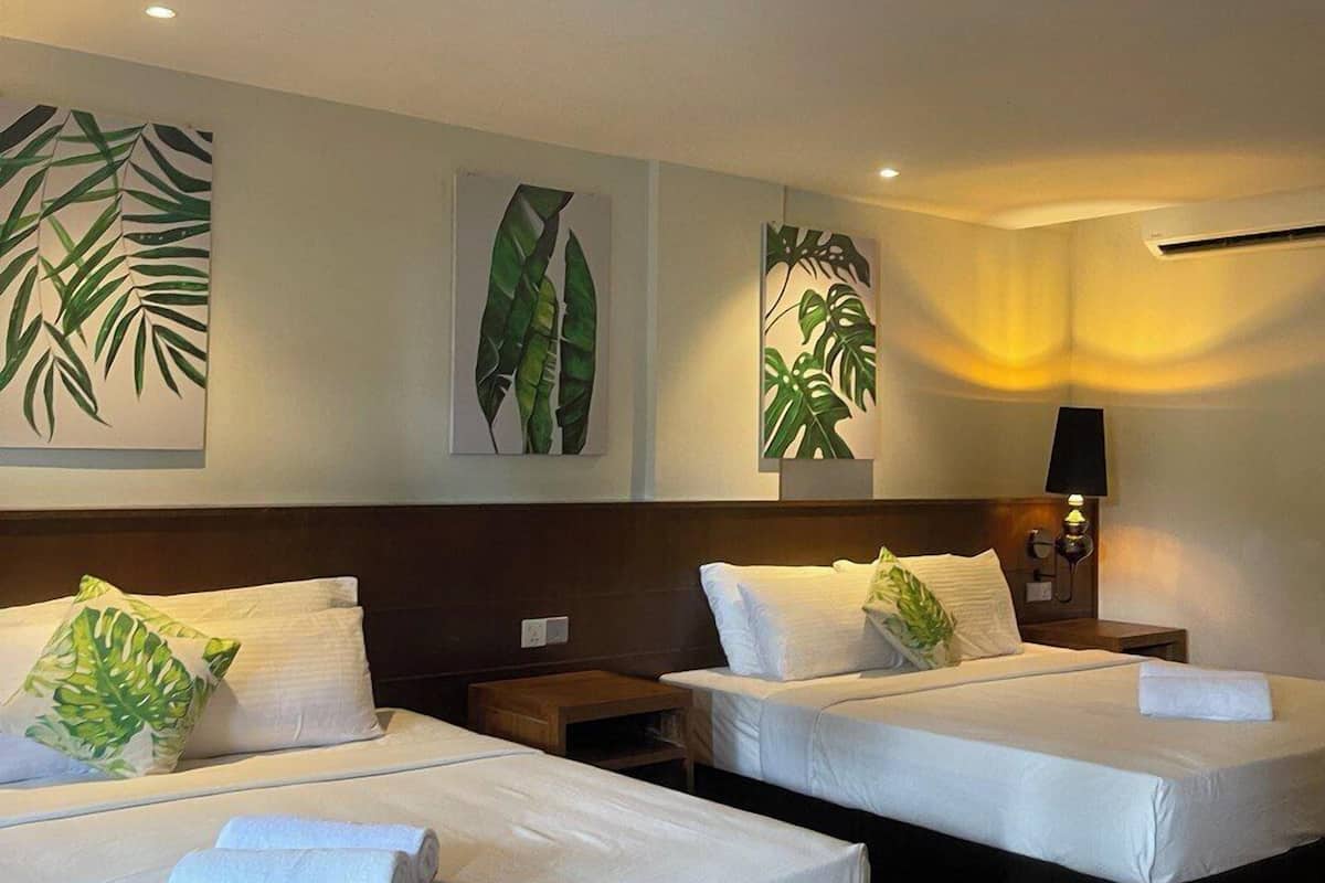 A hotel room with two beds, each with white linens and green leaf-patterned pillows. There are wall-mounted air conditioning units and a bedside table with a lamp.