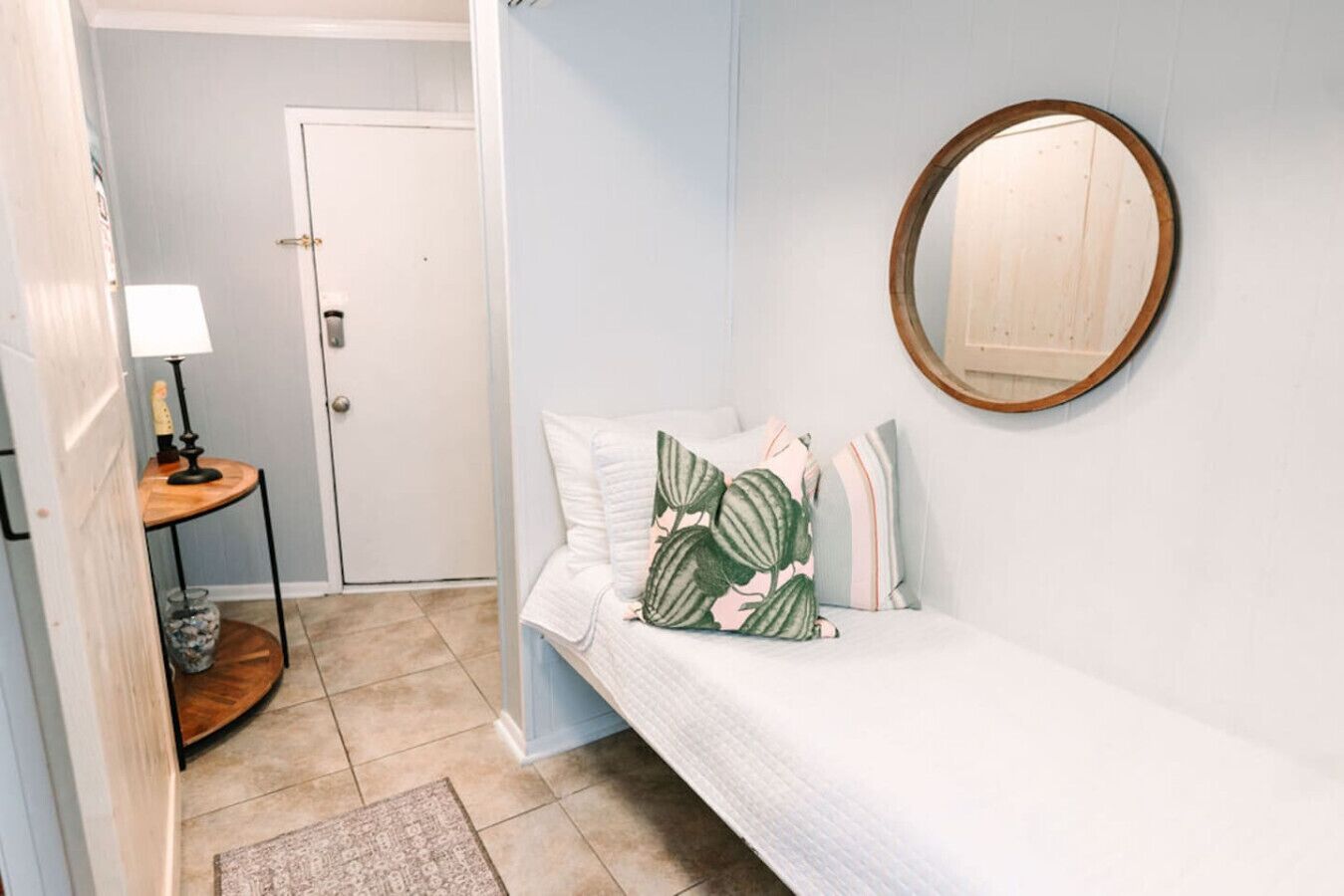 A bedroom with a bed, a round mirror, a bedside table with a lamp, and a door.
