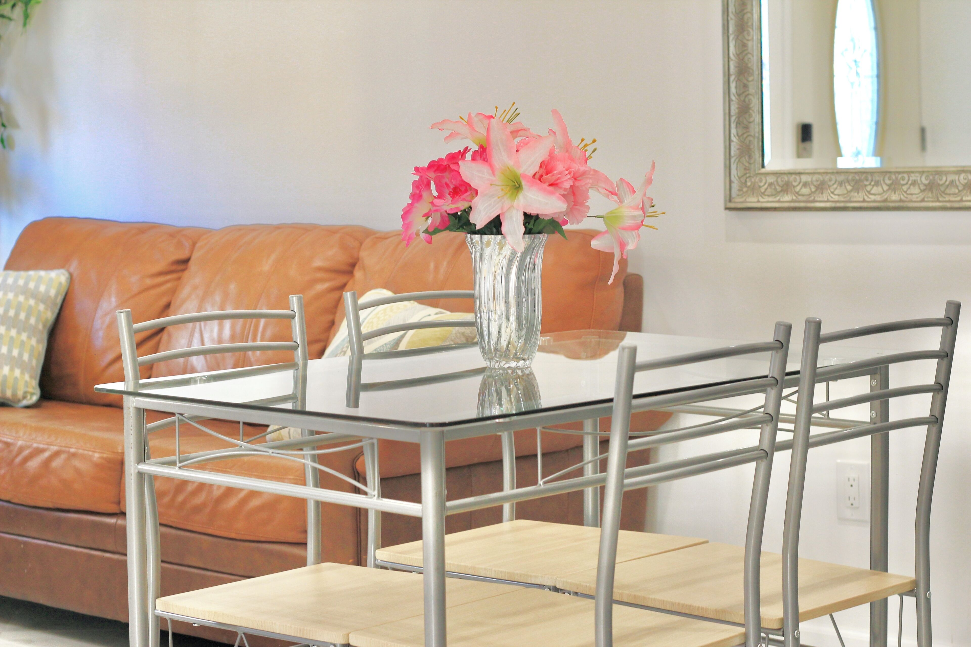 Un comedor con mesa de vidrio y sillas de metal, un jarrón con flores rosadas y un sofá de cuero marrón al fondo.