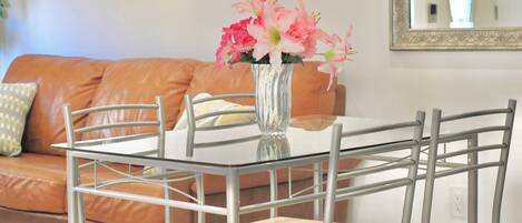 Un comedor con mesa de vidrio y sillas de metal, un jarrón con flores rosadas y un sofá de cuero marrón al fondo.