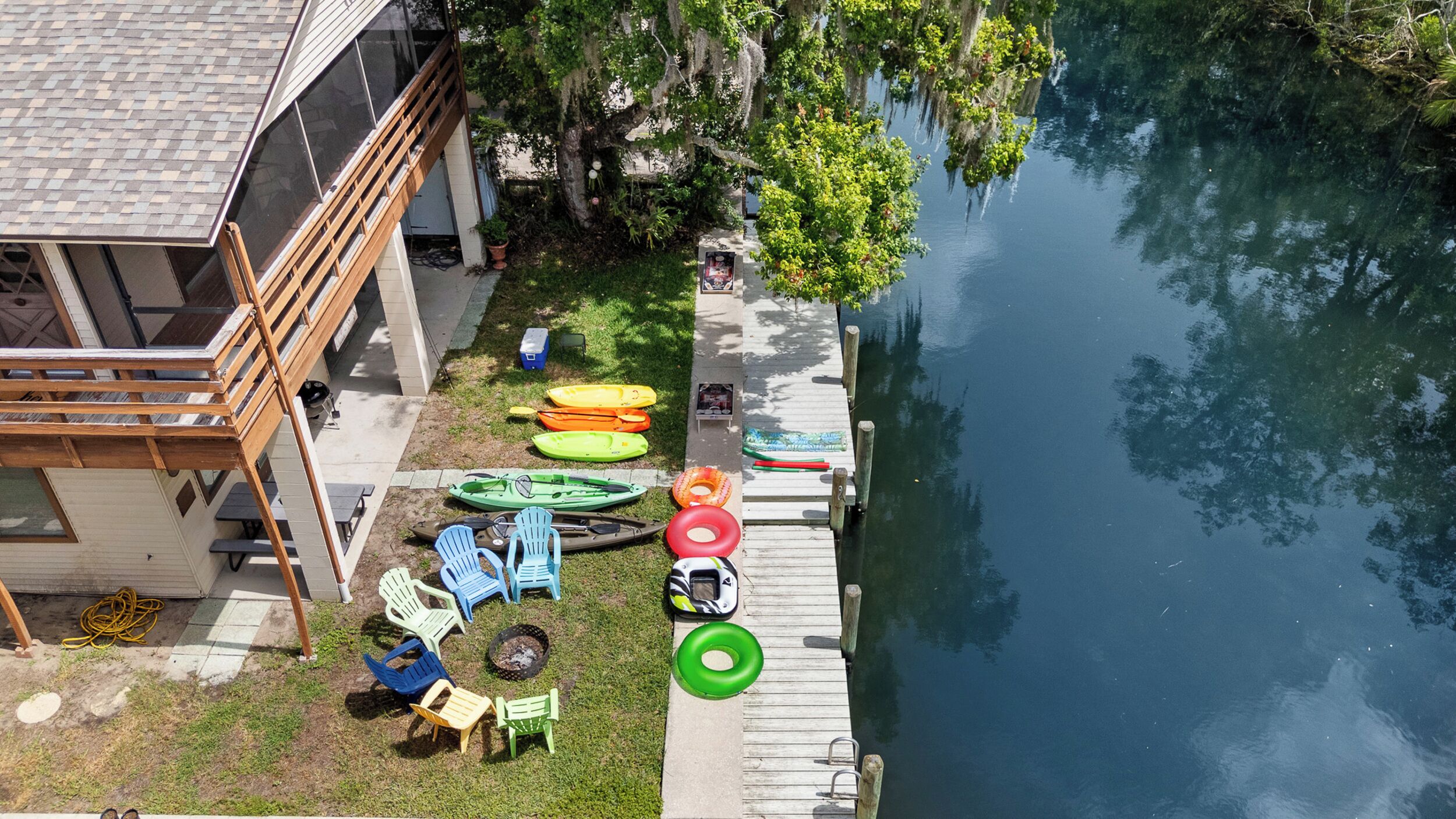 Un muelle con kayaks y una tabla de remo, un césped con sillas Adirondack, y un edificio con tejado inclinado.