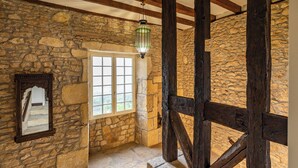 Una habitación con paredes de piedra y estructura de vigas de madera, una ventana con contraventanas blancas y un farol colgante.